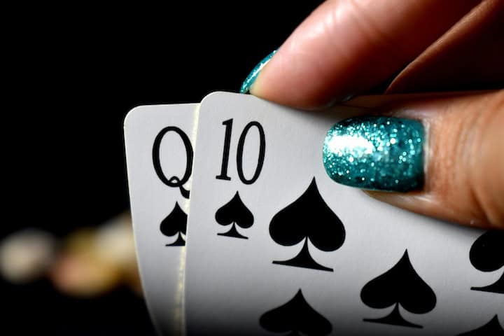 A hand with glittering green fingernails turns up the corner of two playing cards to show a Queen and 10 of spades.