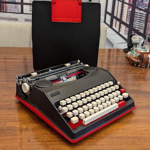 A black manual typewriter from the 80s with white keys and a red base. Behind it sits its carrying case in black and red. It's very Batmobile-coded.