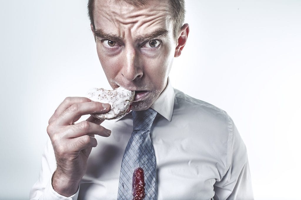 A businessman with a short haircut, bushy eyebrows, and a furrowed forehead stares into the camera while biting into a powdered donut. A dollop of jelly has dripped onto his tie.