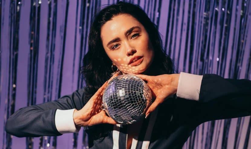 A dark-haired woman in a dark blue jumpsuit with white trim holds a small disco ball in her hands. A purple curtain is behind her.