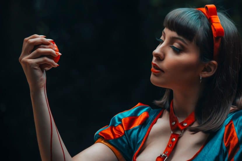 A woman dressed as Snow White stands in darkness, clutching an apple. A rivulet of red runs from her hand down her forearm.