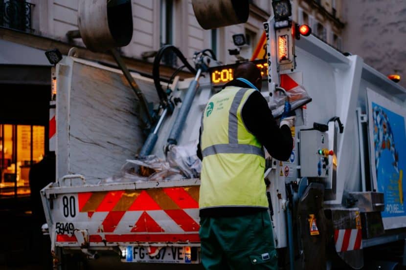 A worker in a yellow high-visibility vest stands at the back of a garbage truck on a busy city street