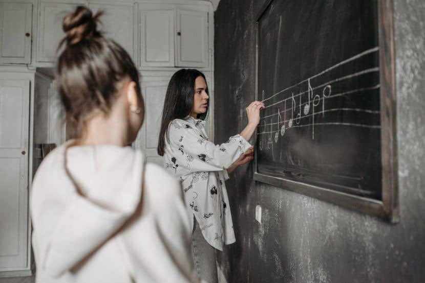 A woman in a grey shirt stands at a chalkboard writing out a musical scale while a young woman in a white hoodie watches on.